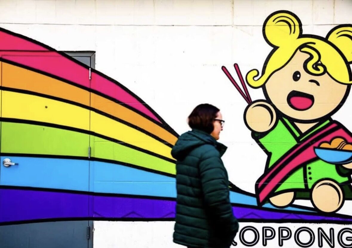 Sushi mural on the side of a sushi restaurant. It's a girl with chopsticks in her hand. A woman is walking by the mural.